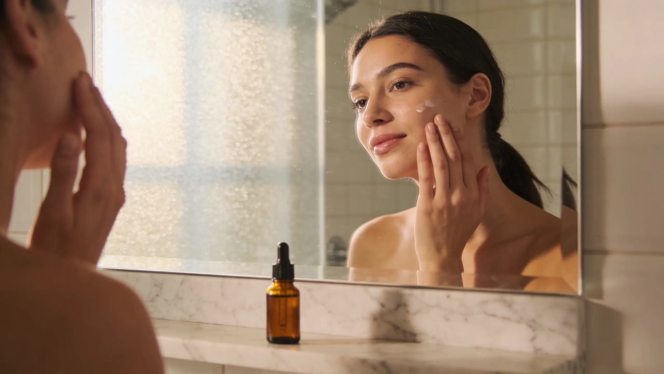 Woman applying serum to her face in front of a bathroom mirror with an amber dropper bottle on a marble ledge